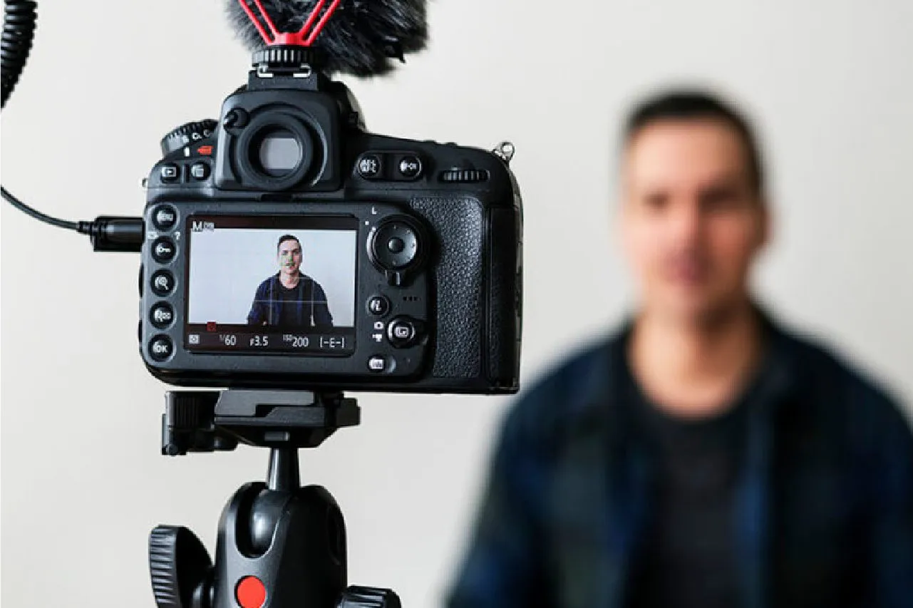DSLR camera on tripod recording a man speaking in front of a blurred background.